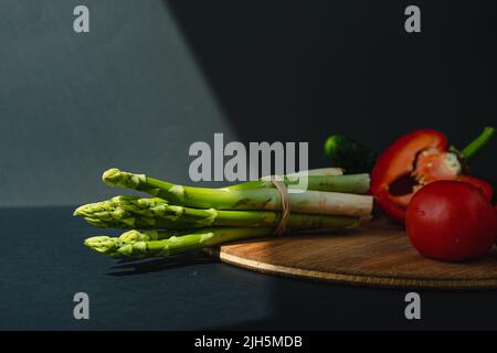 rami di asparagi verdi freschi, peperoni e pomodori su un asse di legno, sfondo grigio scuro, vista dall'alto. Concetto di tendenza di base con spazio di copia Foto Stock