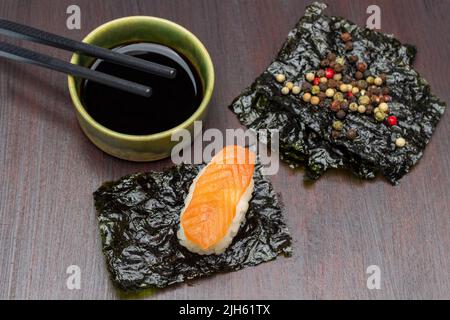 Sushi con salmone su foglio nori. Allspice su foglio nori. Salsa di soia e bacchette in ciotola. Vista dall'alto. Sfondo marrone. Foto Stock