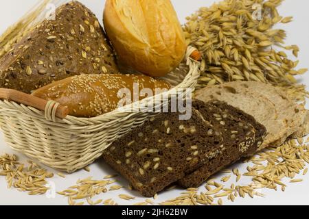 Diversi tipi di pane: Bianco e nero con semi, baguette e panini in un cestino di vimini. Grani di avena e rametti di avena Foto Stock