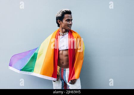 Bell'uomo muscoloso che si posa contro la parete grigia con bandiera arcobaleno Foto Stock