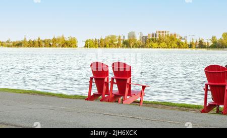 Due sedie rosse Adirondack sull'erba verde di un molo. Vista sul grande lago blu con un cielo blu perfetto. Concetto di luogo ideale di riposo. Dow's l Foto Stock