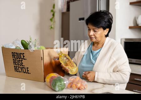 Donna matura biraciale con capelli corti che disimpaccano i generi alimentari dalla scatola riutilizzabile sul tavolo a casa Foto Stock