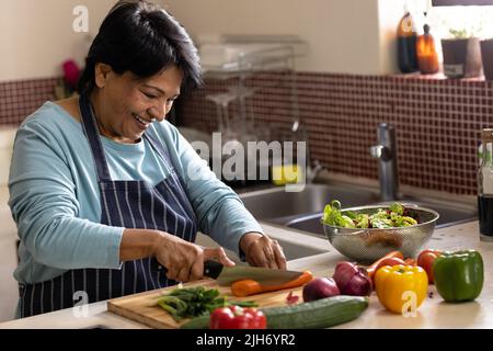 Donna matura sorridente biraciale con capelli corti indossando grembiule tagliando verdure a bordo in cucina Foto Stock