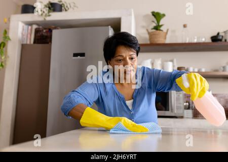Vista ad angolo basso di donna matura biraciale indossando guanti pulizia isola cucina con straccio, spazio copia Foto Stock