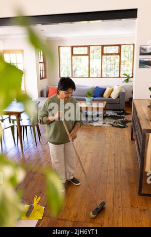 Vista ad angolo alto di donna matura biraciale con capelli corti che spazzano pavimento in legno duro con scopa a casa Foto Stock