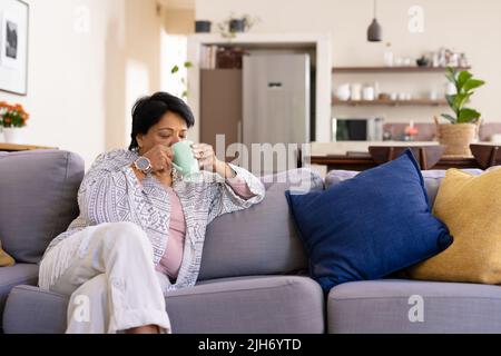 Donna matura biraciale con capelli corti che bevono caffè mentre si rilassa sul divano in salotto Foto Stock