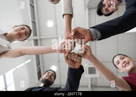 Concetto di team building. I colleghi si stanno in piedi in cerchio battendo il pugno insieme, celebrando il successo di affari in ufficio moderno con alto soffitto. Vie inferiore Foto Stock