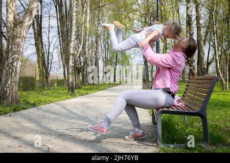 Su una panchina in un parco cittadino, una madre solleva il suo bambino in alto nell'aria. Foto Stock