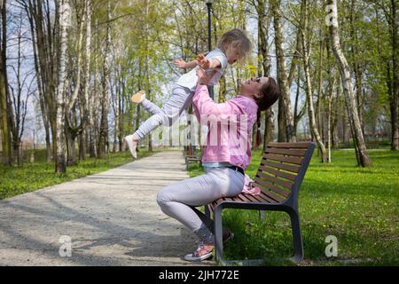 Su una panchina in un parco cittadino, una madre solleva il suo bambino in alto nell'aria. Foto Stock