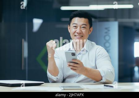 Successo vincente. Il giovane uomo d'affari asiatico tiene il telefono in mano, gioisce vincere, buone notizie, legge il messaggio. Sorridente. Si siede in un ufficio moderno a un tavolo con un notebook e un tablet, guardando la fotocamera Foto Stock