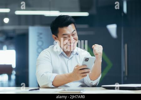 Successo vincente. Il giovane uomo d'affari asiatico tiene il telefono in mano, gioisce vincere, buone notizie, legge il messaggio. Sorridente. Si siede in un ufficio moderno a un tavolo con un notebook e un tablet Foto Stock