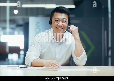 Pullman per affari. Un giovane uomo asiatico in cuffie con microfono conduce l'allenamento professionale con la fotocamera, spiega, dice, sorride. Seduto dietro una scrivania in un ufficio moderno. Foto Stock