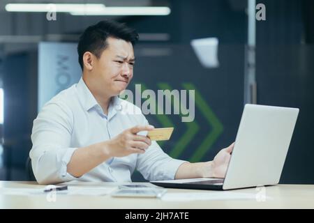 Preoccupato giovane uomo d'affari asiatico che guarda sorpreso a computer portatile e carta di credito in mano. Seduto in ufficio alla scrivania Foto Stock