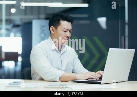 Serio e concentrato giovane uomo d'affari asiatico, libero professionista, imprenditore. Sedersi a un tavolo in un ufficio moderno, lavorare con un computer portatile e documenti. Foto Stock
