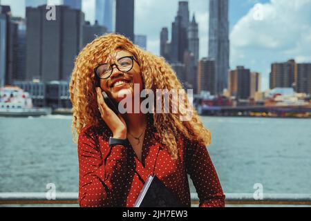 Ritratto giovane imprenditore adulto donna millennial con occhiali e capelli afro sorridente parlare al telefono all'aperto con Manhattan New York City skyline dietro il fiume Hudson Foto Stock