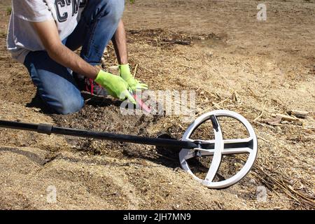 Un ragazzo cammina lungo la spiaggia alla ricerca di tesori con un metal detector wireless in mano e un puntatore nell'altra mano. Alberi nel backgrou Foto Stock