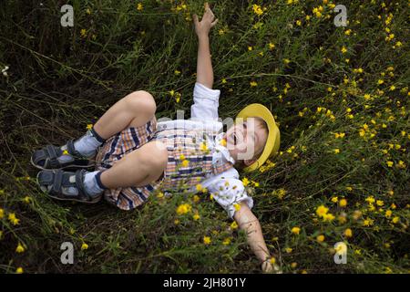 carino ragazzo allegro in un cappello giallo si trova su un campo tra fiori gialli selvatici. Umore estivo, spensierata infanzia gioiosa, coccole. Esplora la natura in vacanza Foto Stock