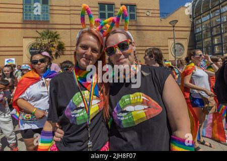 Southend on Sea, Regno Unito. 16 luglio 2022. Due persone si mettono insieme a una celebrazione dell'orgoglio. Entrambi indossano t-shirt nere abbinate con labbra arcobaleno e la parola "ORGOGLIO", accessoriate con occhiali da sole, sciarpe, braccialetti e orecchie coniglietti a tema arcobaleno. Lo sfondo mostra una folla vivace vestita con bandiere arcobaleno e abbigliamento festivo, celebrando l'orgoglio e la solidarietà LGBTQ+. 1000 partecipanti si riuniscono per l'evento Southend Pride, prima che la parata inizi lungo High Street e nei Warrior Square Gardens per un Pride Festival con spettacoli dal vivo. Penelope Barritt/Alamy Live News Foto Stock