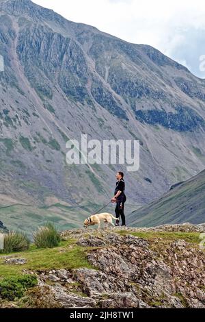 Un uomo di mezza età con il cane in piedi su un affioramento roccioso con la Grande montagna Gable sullo sfondo ammirando la vista in Wasdale Cumbria Inghilterra Regno Unito Foto Stock