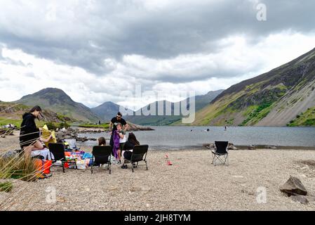 Un grande gruppo di famiglie si diverta una giornata seduti sulla riva del lago di Wastwater con il suggestivo paesaggio montano in lontananza Wasdale Cumbria UK Foto Stock