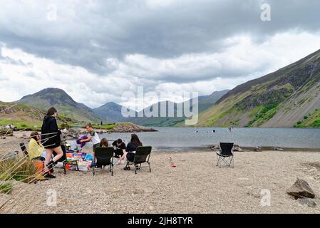 Un grande gruppo di famiglie si diverta una giornata seduti sulla riva del lago di Wastwater con il suggestivo paesaggio montano in lontananza Wasdale Cumbria UK Foto Stock