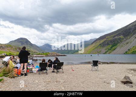 Un grande gruppo di famiglie si diverta una giornata seduti sulla riva del lago di Wastwater con il suggestivo paesaggio montano in lontananza Wasdale Cumbria UK Foto Stock