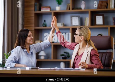 Due giovani donne di affari sorridenti che danno high-five in ufficio moderno. Buone lavoratrici che festeggiano il successo, buon affare, nuovo contratto, buoni risultati di Foto Stock