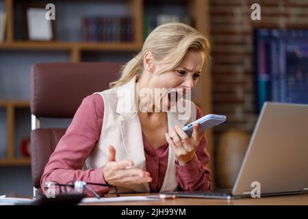 Una donna d'affari furiosa urla allo smart phone mentre risponde alle chiamate in ufficio. Donna arrabbiata infastidita infastidita di cattivo servizio, avendo Foto Stock