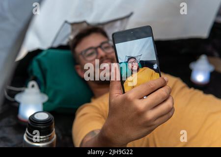 ragazzo in t-shirt gialla e occhiali all'interno della sua tenda in mezzo alla natura tenendo il suo cellulare e prendendo un selfie sdraiato. Foto Stock