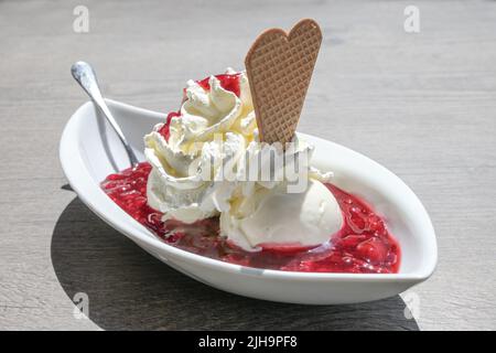 Gelato con panna montata, biscotto al wafer e gelatina di frutta rossa, in Germania chiamata rote gruetze, servito in una ciotola bianca su un tavolo grigio su un summ soleggiato Foto Stock