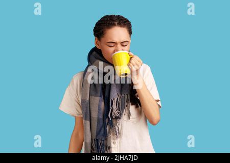 Donna malata dispiaciuta con i doadlocks soffre di tosse e di freddo cattivo, in piedi avvolto in sciarpa, bevande calde, malattia di cure, indossare camicia. Studio interno girato isolato su sfondo blu. Foto Stock