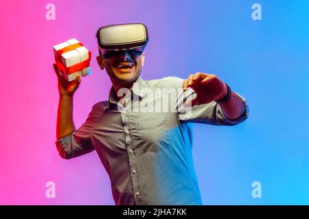 Ritratto di un uomo felice sorridente in camicia con visore vr, che lancia il presente box, dopo aver emozionato l'espressione facciale. Studio interno girato isolato su colorato sfondo di luce al neon. Foto Stock