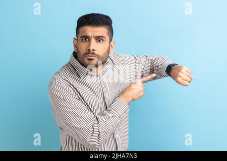 Guarda il tempo. Impaziente uomo d'affari con barba puntata verso l'orologio da polso e aspetto infastidito e dispiaciuto, indossando una camicia a righe. Studio interno girato isolato su sfondo blu. Foto Stock