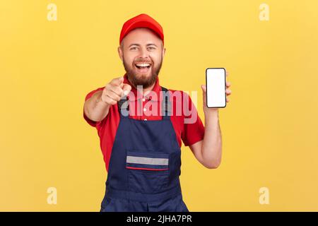 Sorridente uomo lavoratore felice in tuta blu t-shirt rossa puntando il dito contro di te e mostrando smartphone con schermo vuoto, applicazione online per gli ordini di assistenza. Studio interno girato isolato su sfondo giallo Foto Stock