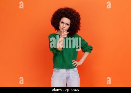 Ritratto di donna rigorosa con acconciatura Afro in piedi con indice puntato dito alla fotocamera che ha serio admonishing espressione facciale. Studio interno girato isolato su sfondo arancione. Foto Stock