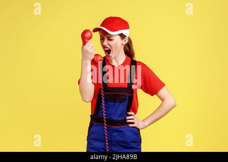 Donna operaia aggressiva urlando al cliente sul telefono, non accetta l'ordine, personale di assistenza incompetente, indossare tute e cappuccio rosso. Studio interno girato isolato su sfondo giallo. Foto Stock