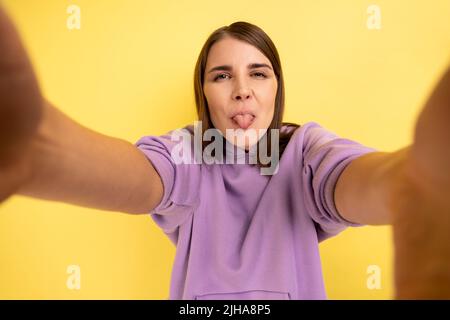 Giovane adulta bambino attraente donna guardando la macchina fotografica con espressione divertente e mostrando lingua fuori POV, indossando la felpa con cappuccio viola. Studio interno girato isolato su sfondo giallo. Foto Stock