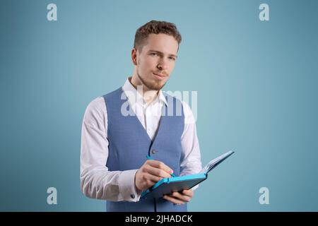 Uomo d'affari che tiene il notepad per la scrittura nel vestito su sfondo blu in studio. Concetto di pianificazione Foto Stock