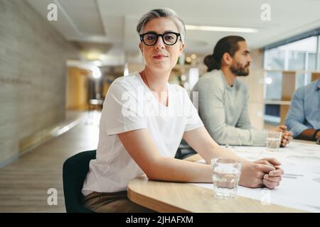 Professionista di design maturo che guarda la fotocamera mentre si siede in una riunione con il suo team. Gruppo di imprenditori innovativi che si sfidano mentre lavorano Foto Stock