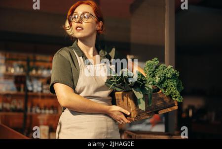 Donna proprietario negozio di alimentari guardando via pensieroso mentre tiene una scatola di verdure fresche biologiche nel suo negozio. Giovane imprenditore femminile che gestisce un Foto Stock