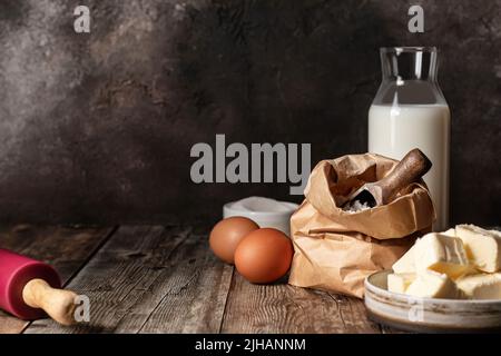 Ingredienti di cottura: Latte, farina, uova di burro e spilla su sfondo di legno. Tasto scuro, stile rustico, spazio di testo. Orientamento orisontale Foto Stock