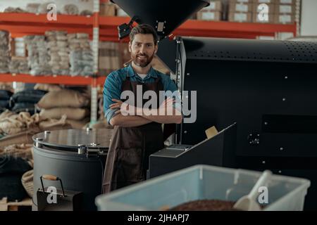 Il proprietario di affari sorridente sta in piedi sullo sfondo della macchina di tostatura del caffè Foto Stock