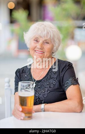 Donna matura seduta a un tavolo in un bar estivo e bere birra Foto Stock