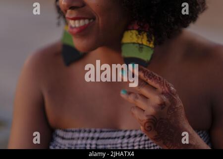 Sorridente Donna nera con capelli afro tiene grande Rastafarian orecchino. Mano con tatuaggio all'henné e unghie all'acqua. Parte superiore del tubo in pied de poule. Chiudere il ritaglio della faccia. Foto Stock