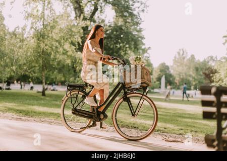 Bella giovane donna in bicicletta elettrica con fiori nel cesto Foto Stock