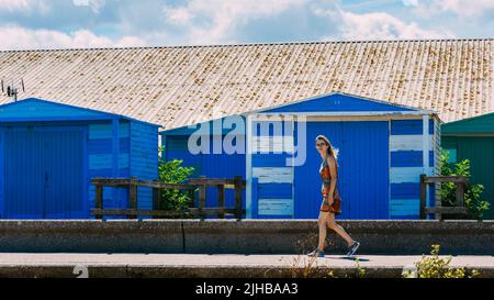 La giovane donna cammina accanto a una capanna di spiaggia blu a Whitstable, Kent, Regno Unito Foto Stock