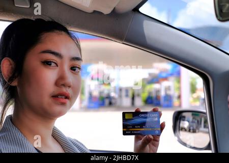 Felice bella donna asiatica seduta all'interno della sua auto mostrando il pagamento della carta di credito in una stazione di servizio Foto Stock