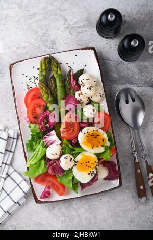 sparagus, pomodoro, lattuga, mozzarella, sesamo nero, lino, insalata di olio d'oliva e uovo bollito morbido su piastra rettangolare in ceramica su tavola grigia in calcestruzzo bac Foto Stock