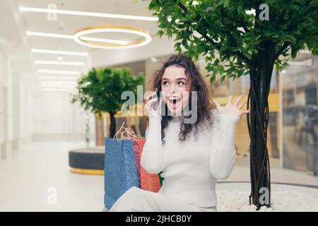 Felice e soddisfatto giovane bella ragazza curly, donna seduta su una panchina nel centro commerciale. Tiene un telefono in mano, chiama, scrive messaggi, condivide le sue impressioni di shopping. Sorride Foto Stock