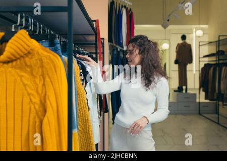 Giovane bella donna con capelli ricci sceglie, alla ricerca di una giacca bianca in un negozio di abbigliamento, la esamina su un appendiabiti sulla rastrelliera. Vuole comprare, sorride Foto Stock